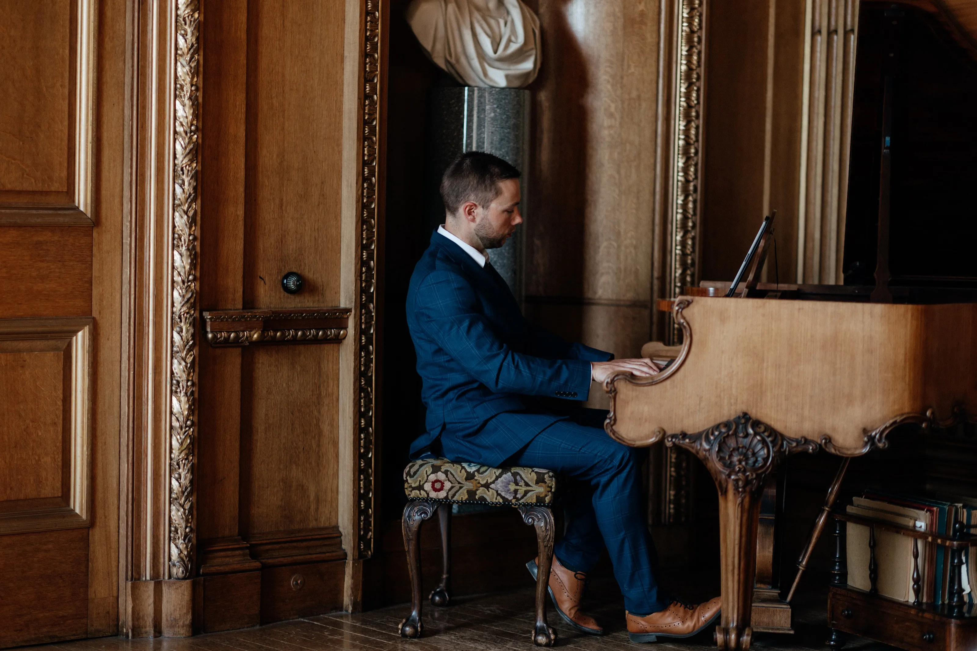 Finn in a navy suit playing an ornate walnut grand piano in an oak-panelled room